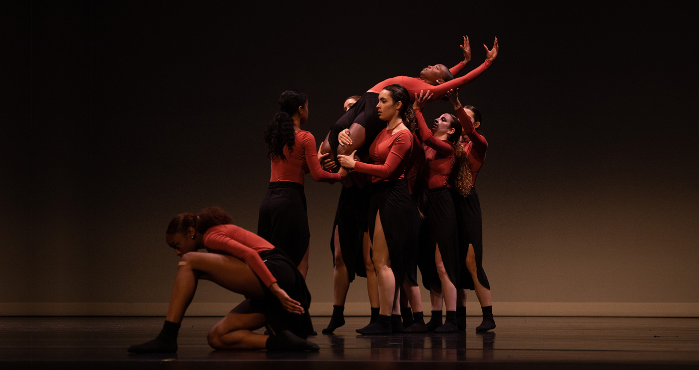 Six dancers in red and brown performing on a brown-lit stage; three women hold one aloft, while another approaches and the last crouch/crawls away
