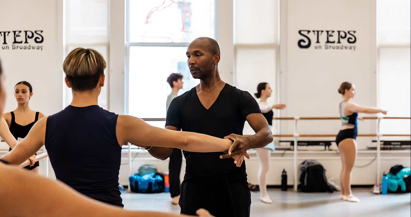 The interior of a dance studio with a teacher helping a young man at the barre