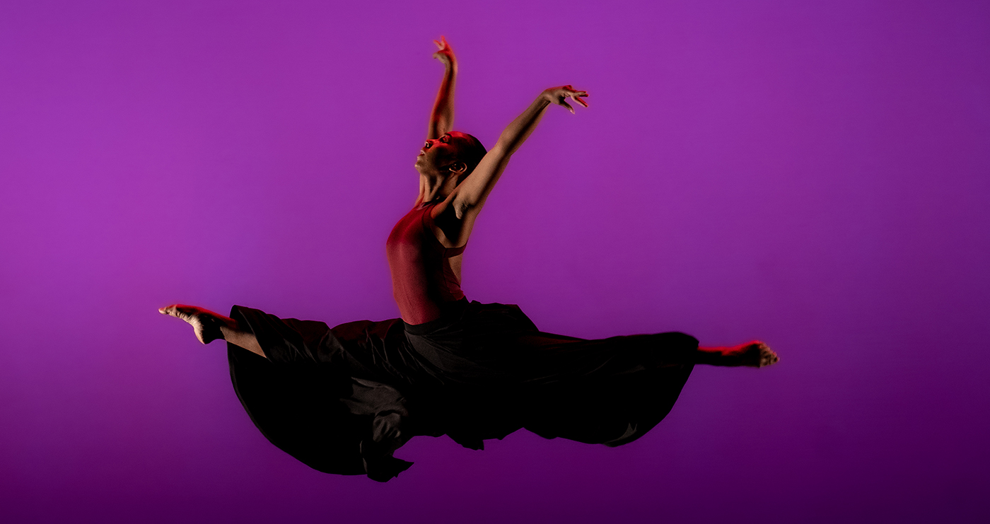 Female dancer in flowing dark skit leaping, arms raised, towards the left of the frame on a stage lit with purple light, she is nearly in silhouette