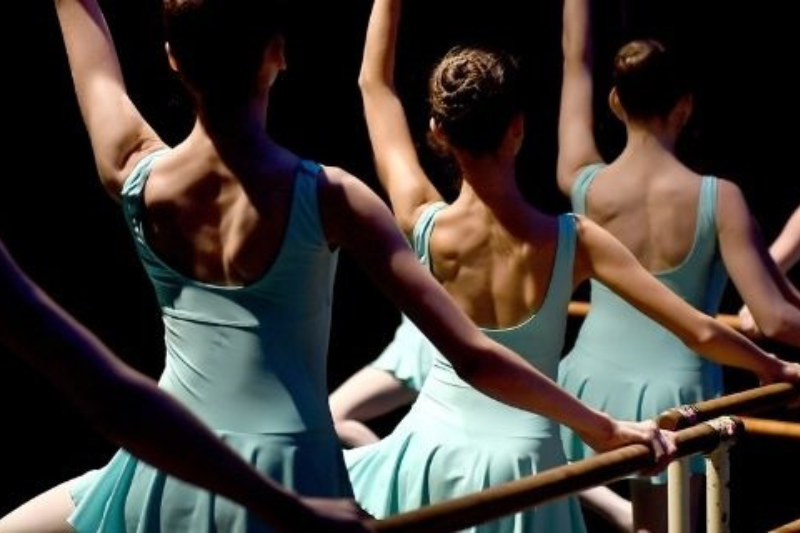 A close up of the backs of three dancers in light blue leotards and skirts arms raised and lit dramatically
